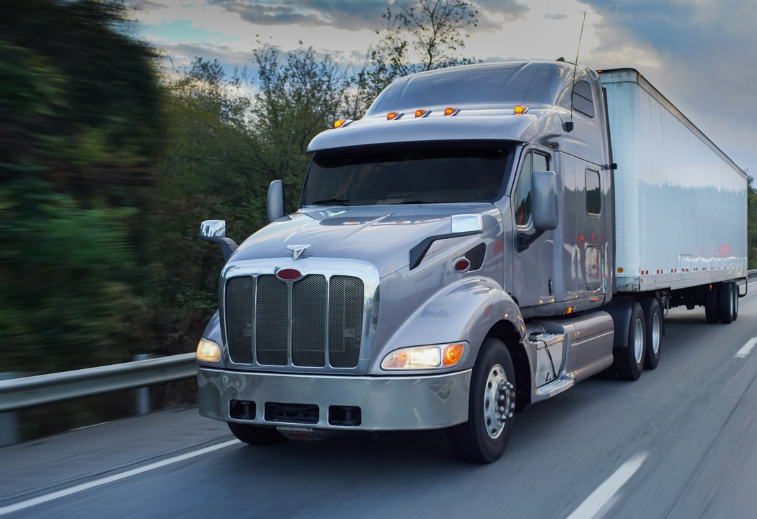 Silver semi truck with a white trailer driving along a highway, with blurred trees and a guardrail in the background