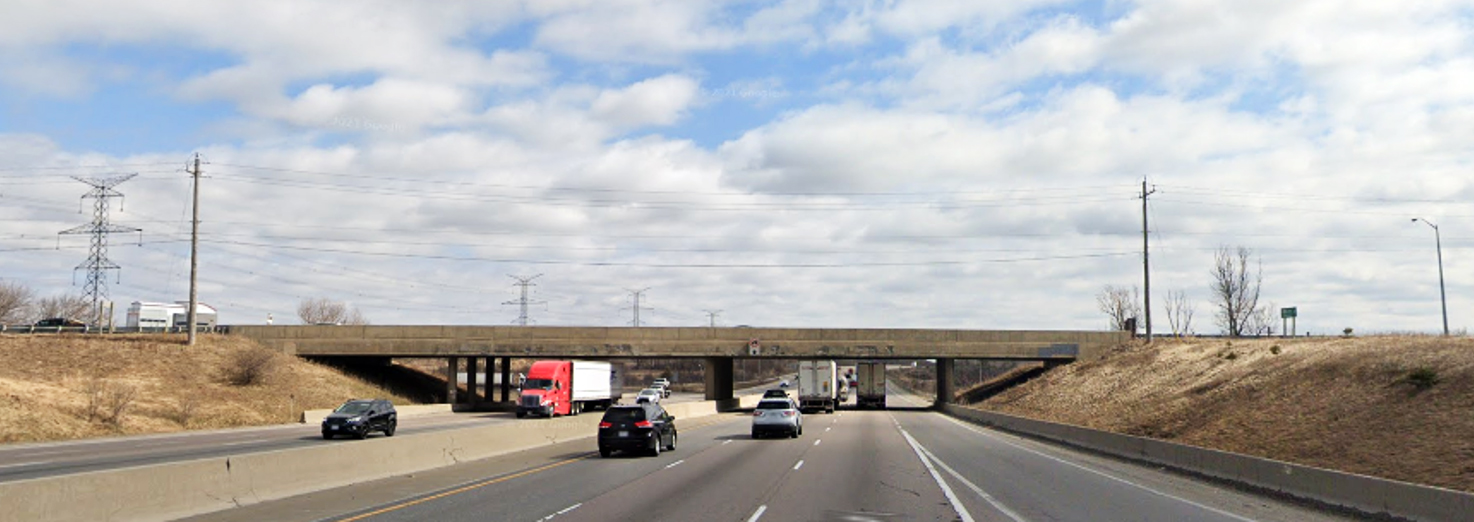 View of Highway 401 traffic passing under the Highway 6 / Brock Road South underpass bridge with grass embankments on either side of the road and a concrete barrier median.